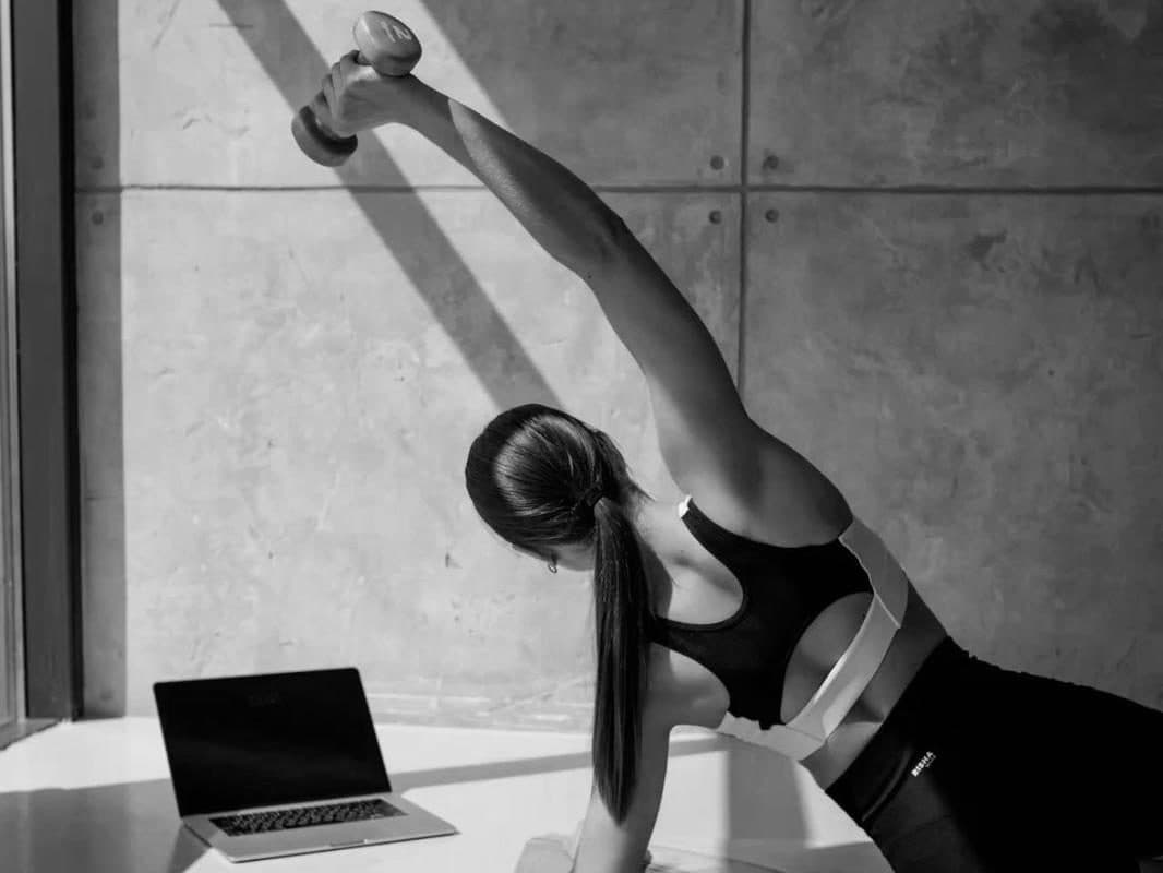 Une femme s'entraîne chez elle avec une haltère, un ordinateur portable ouvert à côté d'elle.