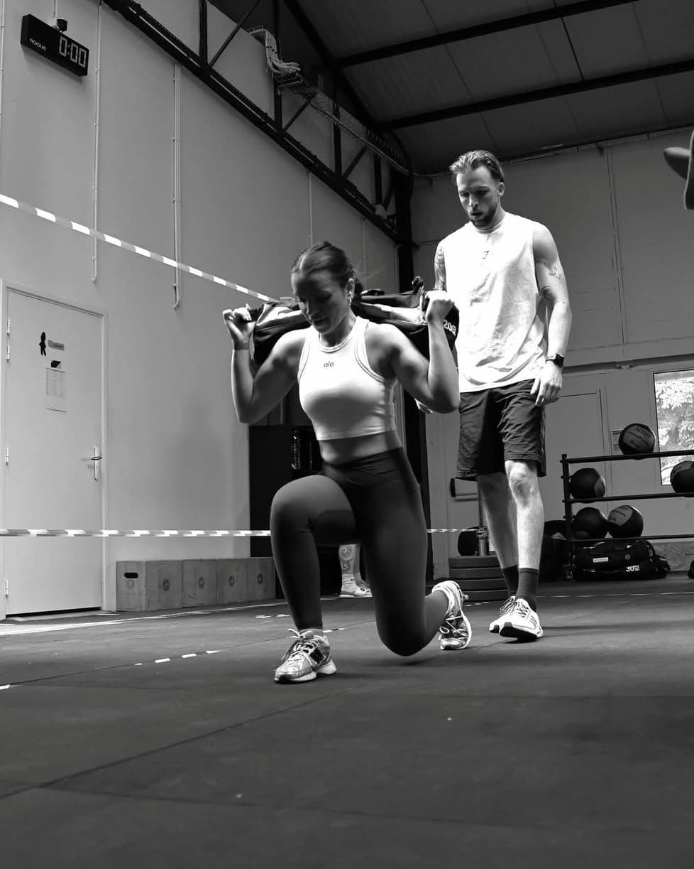 Une coachée en fente avant, sac lesté sur les épaules, corrigée par son coach dans une salle de sport.