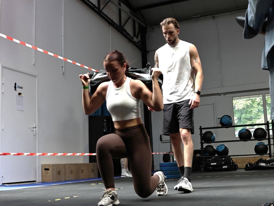 Une coachée en fente avec un sac lesté sur les épaules, un coach derrière elle.
